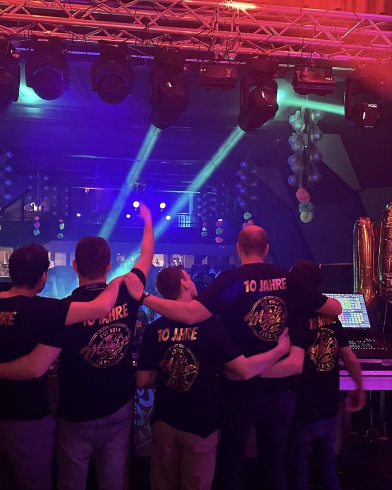 Die Music-Bakery-Crew in „10 Jahre“-Shirts vor der Bühne, über ihnen Moving Heads