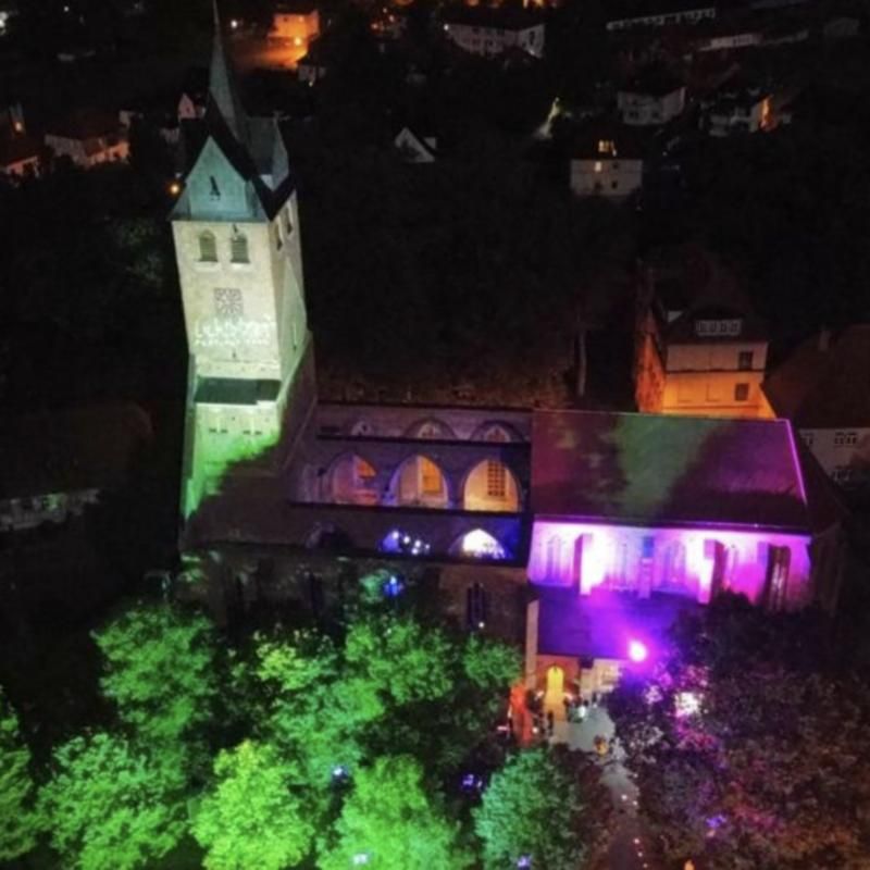 Kirche bei Nacht von oben, Turm und Bäume in Grün, Kirchenschiff in Magenta angestrahlt