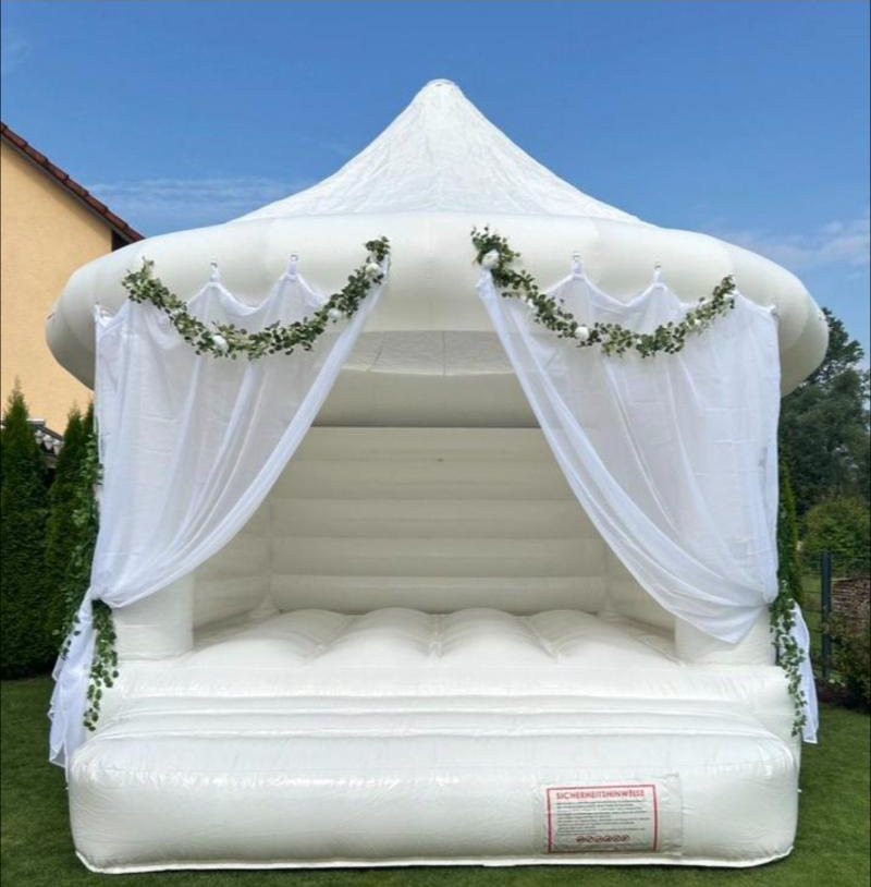 Weiße Hüpfburg White Wedding mit spitzem Dach, weißen Vorhängen und Girlanden aus Grün, aufgebaut auf einer Wiese